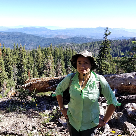 Woman in green long sleeve shirt with bucket hat standing with green trees and mountains in the background