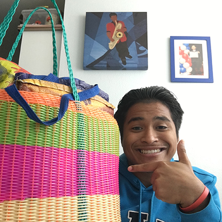 Young teen with black hair wearing a blue sweatshirt smiling and pointing at a colorful woven bag. One painting and a portrait hanging on the wall in the background. 