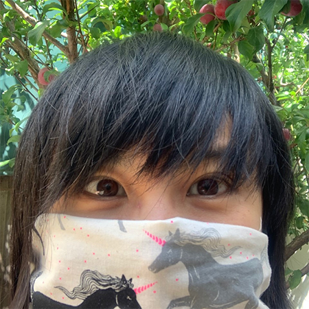 Woman wearing unicorm print face covering with short black hair covering eyebrows in front of fruit tree