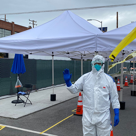 Man in personal protective gear- blue gloves, white bodysuit, blue mask, clear goggles-waving hand standing in front of white tents