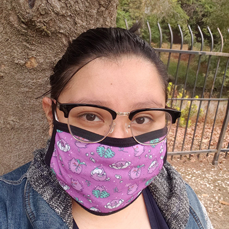 Woman in foreground with black hair wearing black-framed glasses, purple masks with cartoon design, and jean jacket. Tree in background next to gate. 