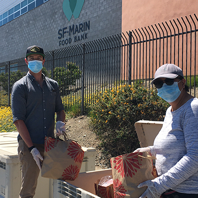 Man wearing cap, mask, and gloves holding paper-bagged groceries with woman wearing baseball cap, mask, glasses, gloves holding bag of groceries