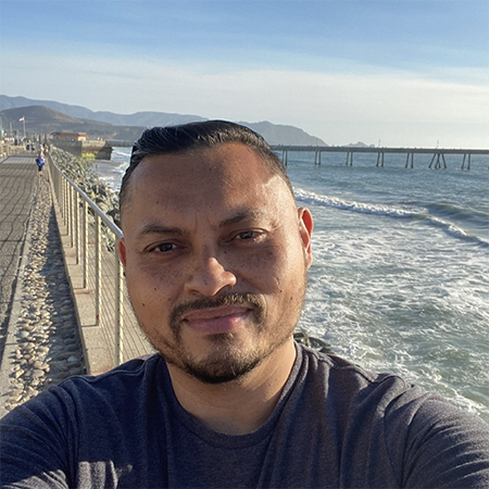 Man with mustache and small beard facing forward with ocean, mountains, and boardwalk in background
