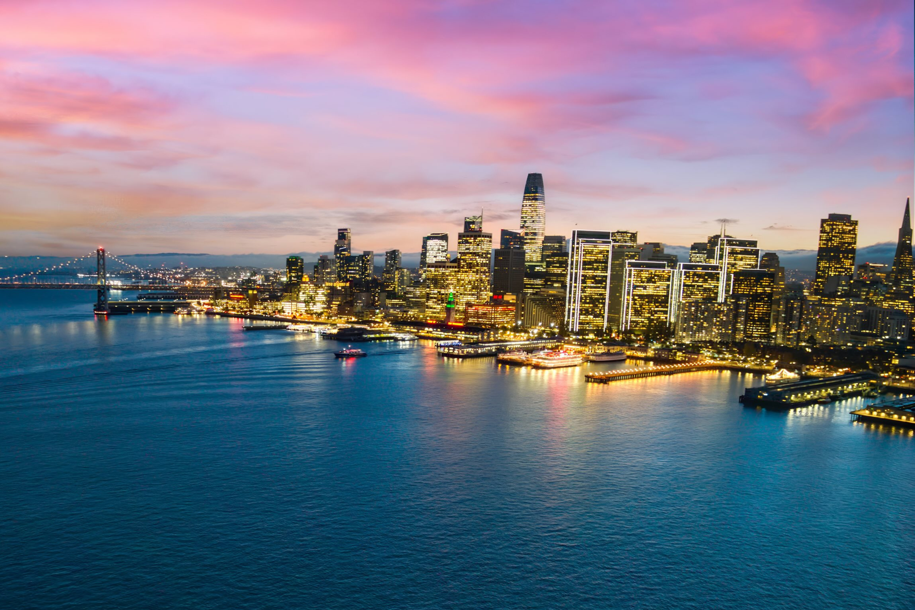 San Francisco skyline at dusk, with the Bay Bridge on the left and illuminated high-rise buildings reflecting on calm bay waters under a pink and purple sunset sky.
