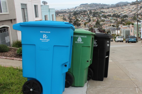 Blue, green, and black waste bins on the street with the city of San Francisco in the background