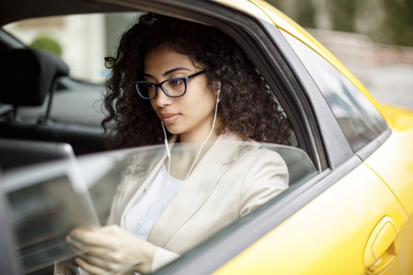A woman with shoulder-length curly black hair and glasses reads a newspaper in the backseat of a taxi car