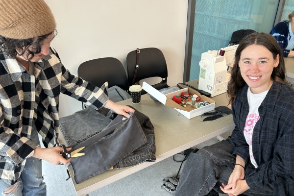 Two people are working on a clothing item with a sewing machine and scissors
