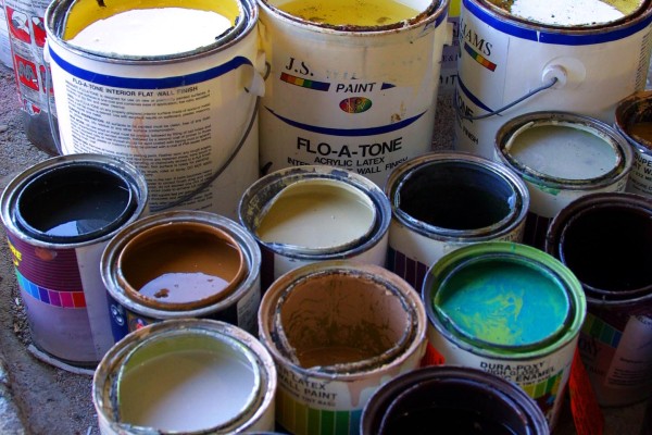 The image shows a collection of paint cans, each filled with different colors of paint. The cans are various sizes and appear to be used, with paint splatters and drips on the rims and sides. The colors include shades of yellow, green, brown, blue, white, and others, indicating a diverse range of paints. The setting looks like a workshop or storage area, with the cans arranged closely together on a surface, likely ready for use in a painting project. The overall scene suggests a practical and creative envir