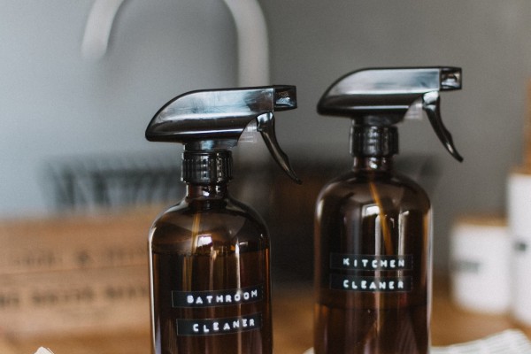 The image shows two brown spray bottles labeled "BATHROOM CLEANER" and "KITCHEN CLEANER," placed on a wooden surface. There is also a wooden dish brush and a white cloth nearby. The setting appears to be a clean and organized kitchen or cleaning area, with a sink and faucet visible in the background. The overall aesthetic is simple and eco-friendly, with a focus on natural materials.