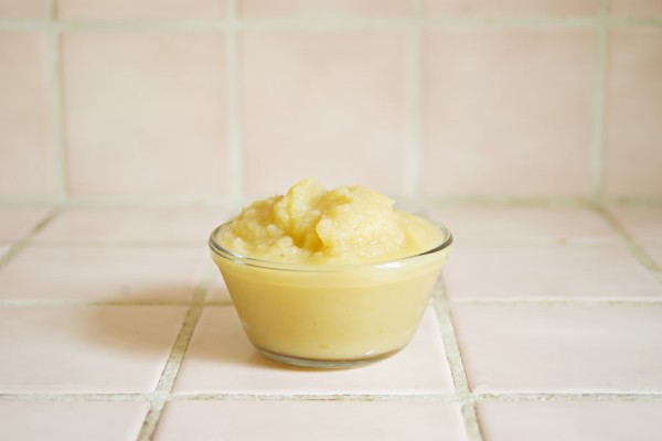 The image shows a small glass bowl filled with a creamy, pale yellow substance, which appears to be applesauce. The bowl is placed on a tiled surface with light pink tiles and white grout. The setting is simple and clean, with the focus on the bowl of applesauce. The overall presentation is neat, and the applesauce looks smooth and freshly made. The soft colors of the tiles complement the light color of the applesauce, creating a visually pleasing and appetizing scene.