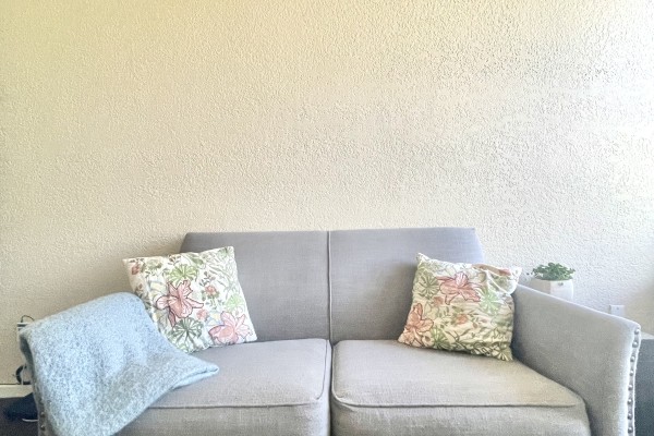 The image shows a grey loveseat with two floral-patterned throw pillows. There is a light blue blanket draped over one arm of the loveseat. The loveseat has a simple design with nailhead trim along the bottom edge and arms. The background is a plain, textured beige wall, and the floor appears to be a dark carpet. To the right of the loveseat, there is a small potted plant on the floor, adding a touch of greenery to the otherwise neutral setting. The overall atmosphere is cozy and inviting, ideal for a relax