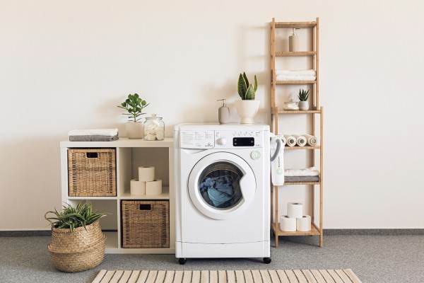 The image shows a well-organized and aesthetically pleasing laundry room. In the center is a front-loading washing machine with some laundry visible inside. To the left of the washing machine, there is a white shelving unit with woven baskets, a small potted plant, a stack of folded towels, and a glass jar filled with laundry items, likely cotton balls or detergent pods. On the floor, there is a woven basket with a green plant.