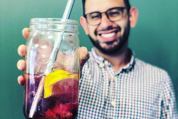 A man with glasses and a checkered shirt is smiling and holding a large mason jar filled with a dark beverage, ice, a metal straw, and a slice of citrus fruit, against a plain green background.