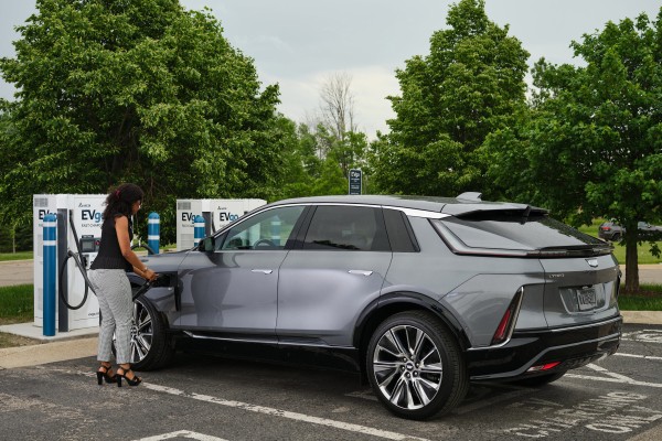 A woman charges a sleek silver electric SUV at an EVgo fast charging station, surrounded by green trees in a parking lot on a cloudy day.