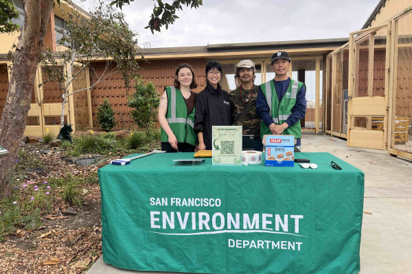 A geren table cover cloth reads SF Environment Department; four volunteers stand behind the table to prepare for an event