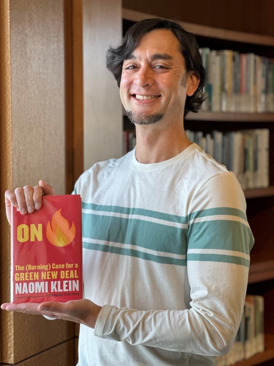 Gregory is standing up holding the book "On Fire" by Naomi Klein. He's smiling and is wearing a white long-sleeved t-shirt.