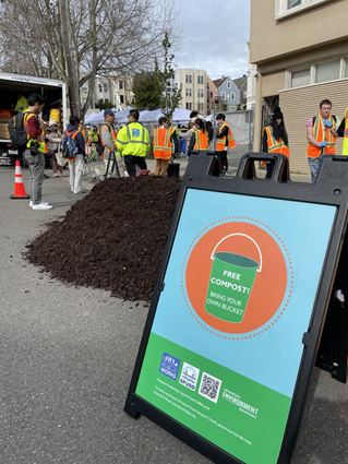 A sign says "Free compost!" with a mound of compost behind the sign. Behind the mound, there is a group of people in line wearing orange vests