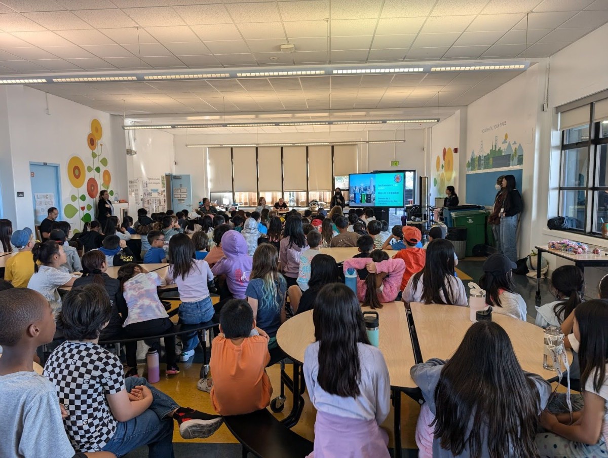 a classroom or event setting with children seated at tables, watching a presentation on a screen