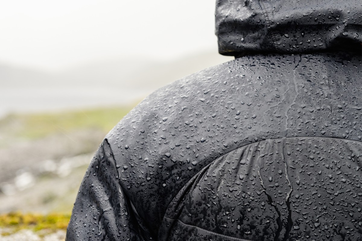 A close-up of a person's shoulder in a water-resistant black jacket. Water droplets are accumulating on the fabric. A blurred, overcast landscape is in the background.