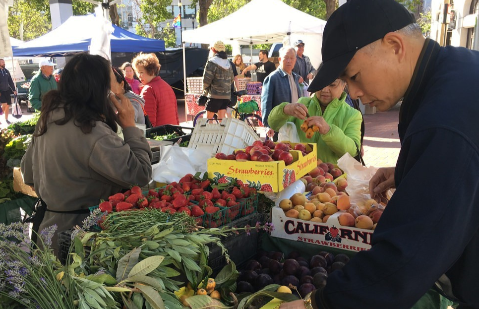 SF resident selecting fresh vegetables at the Heart of the City farmers market. Produce is shown in the background including strawberries, peaches, and plums.