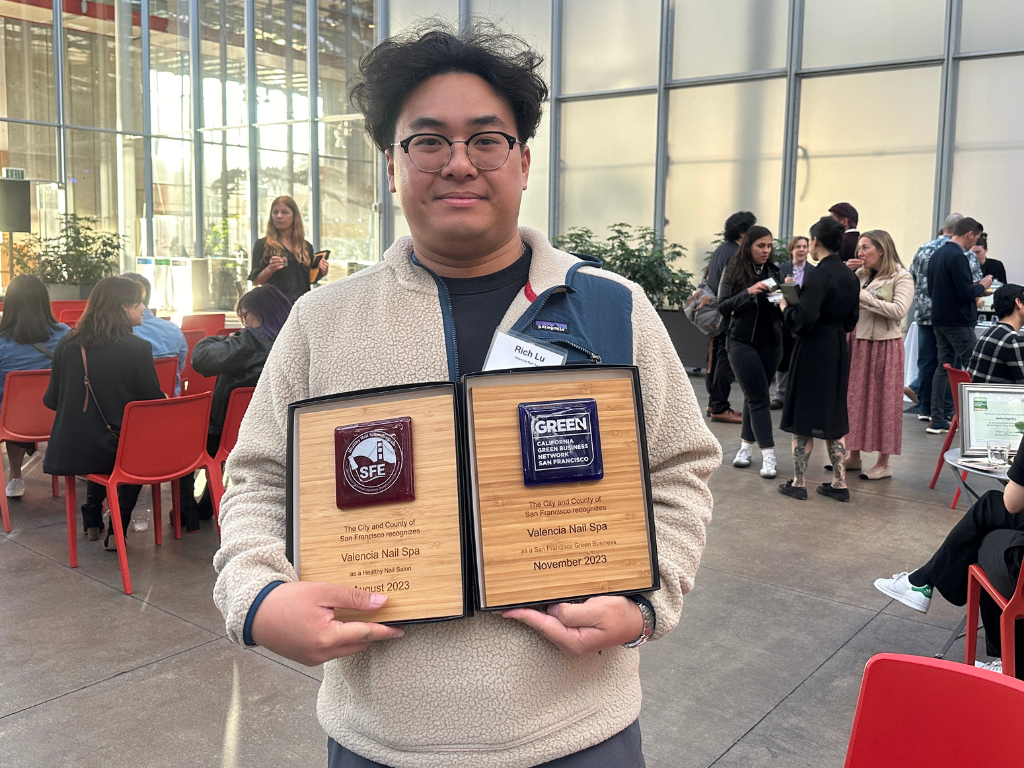 A San Frncisco business owner wearing black rimmed glasses and holding two wooden plaques for environmental awards