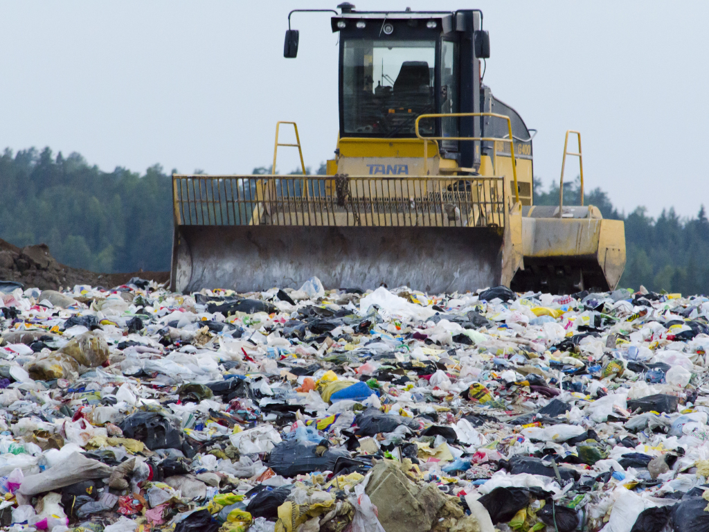 A bulldozer pushing through a large pile of waste at a landfill, with trees visible in the background.