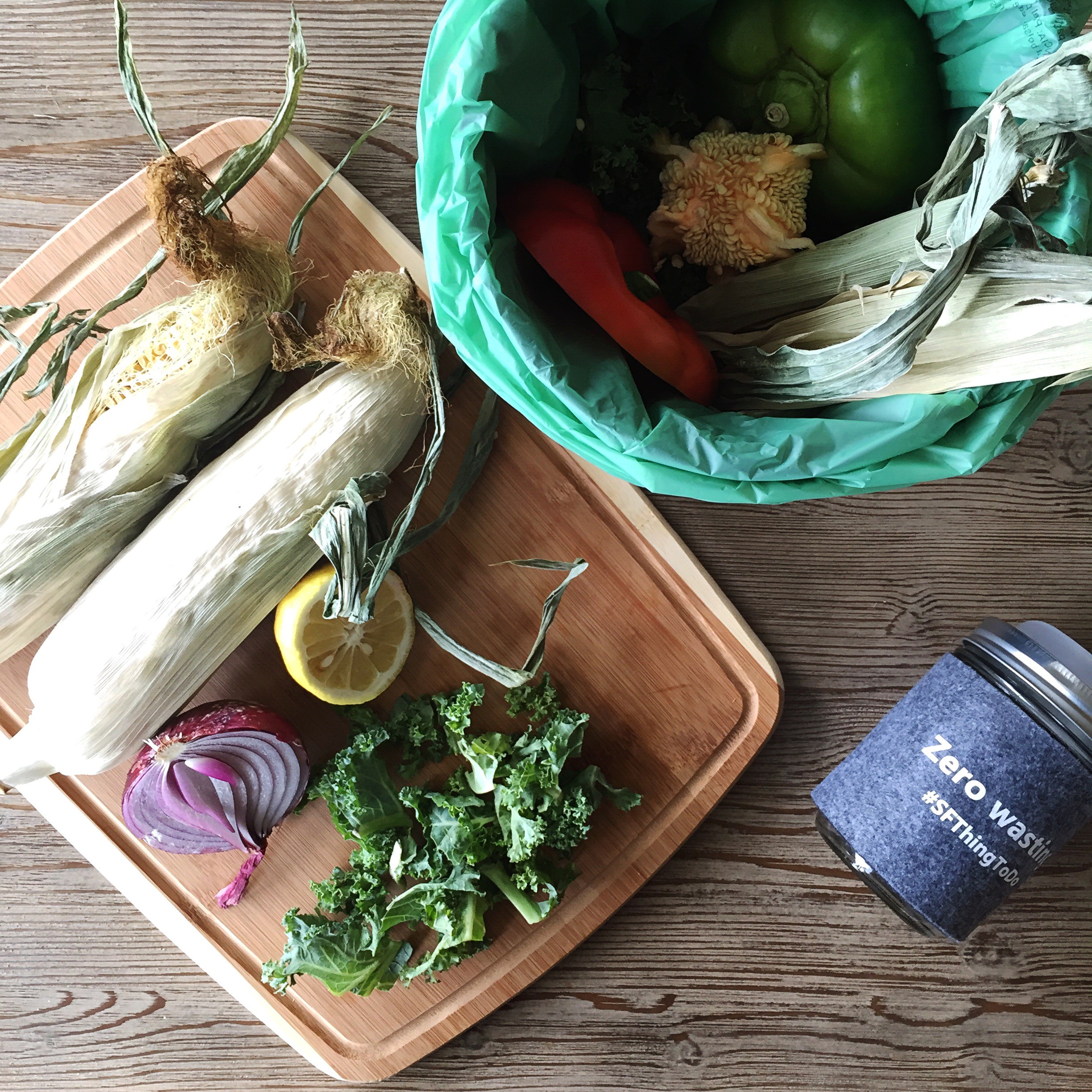 A cutting board with corn husks and kale on top and a compost pail close by for the food scraps
