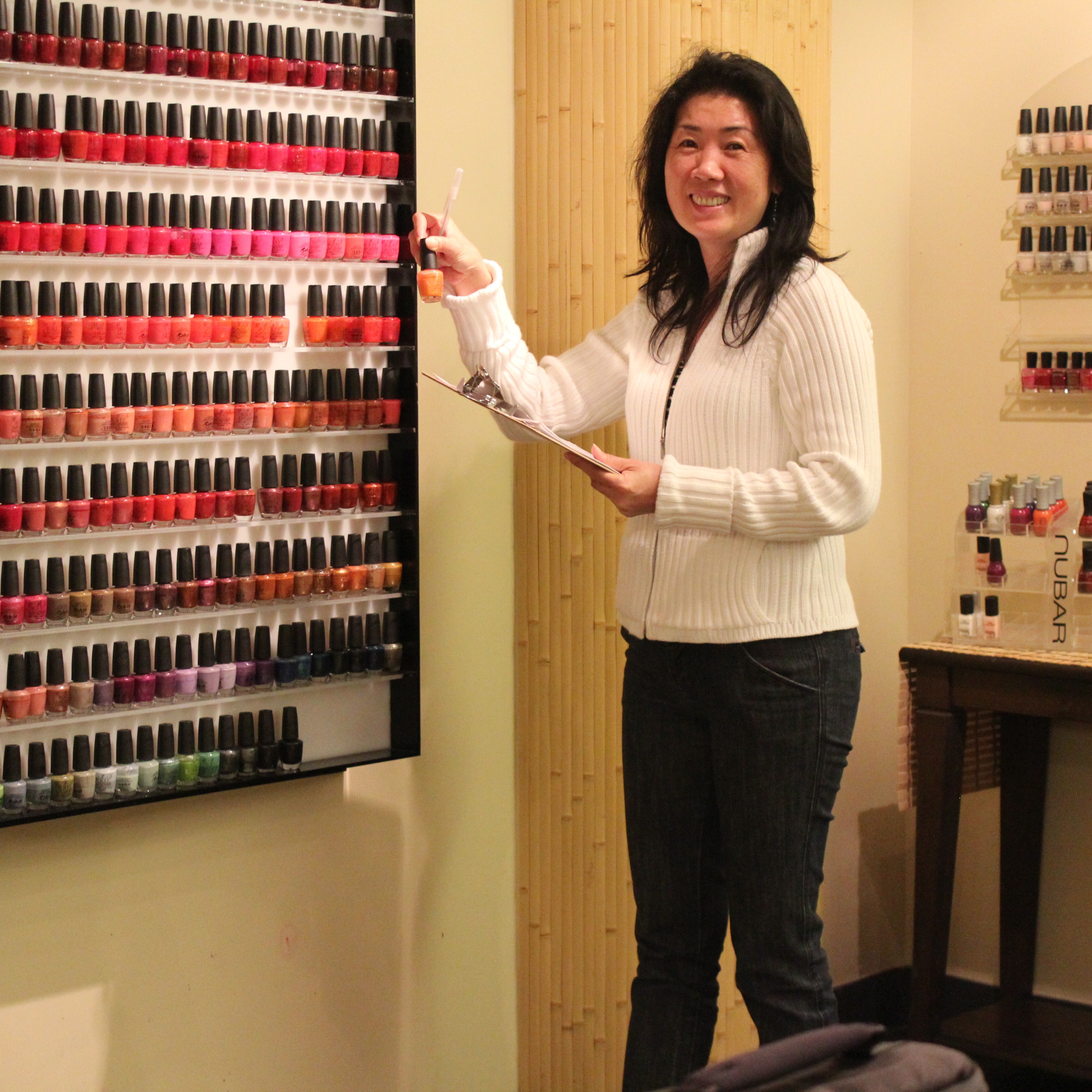 Nail salon owner examines the wall of nail polishes. A lot of them seem to be the color red.