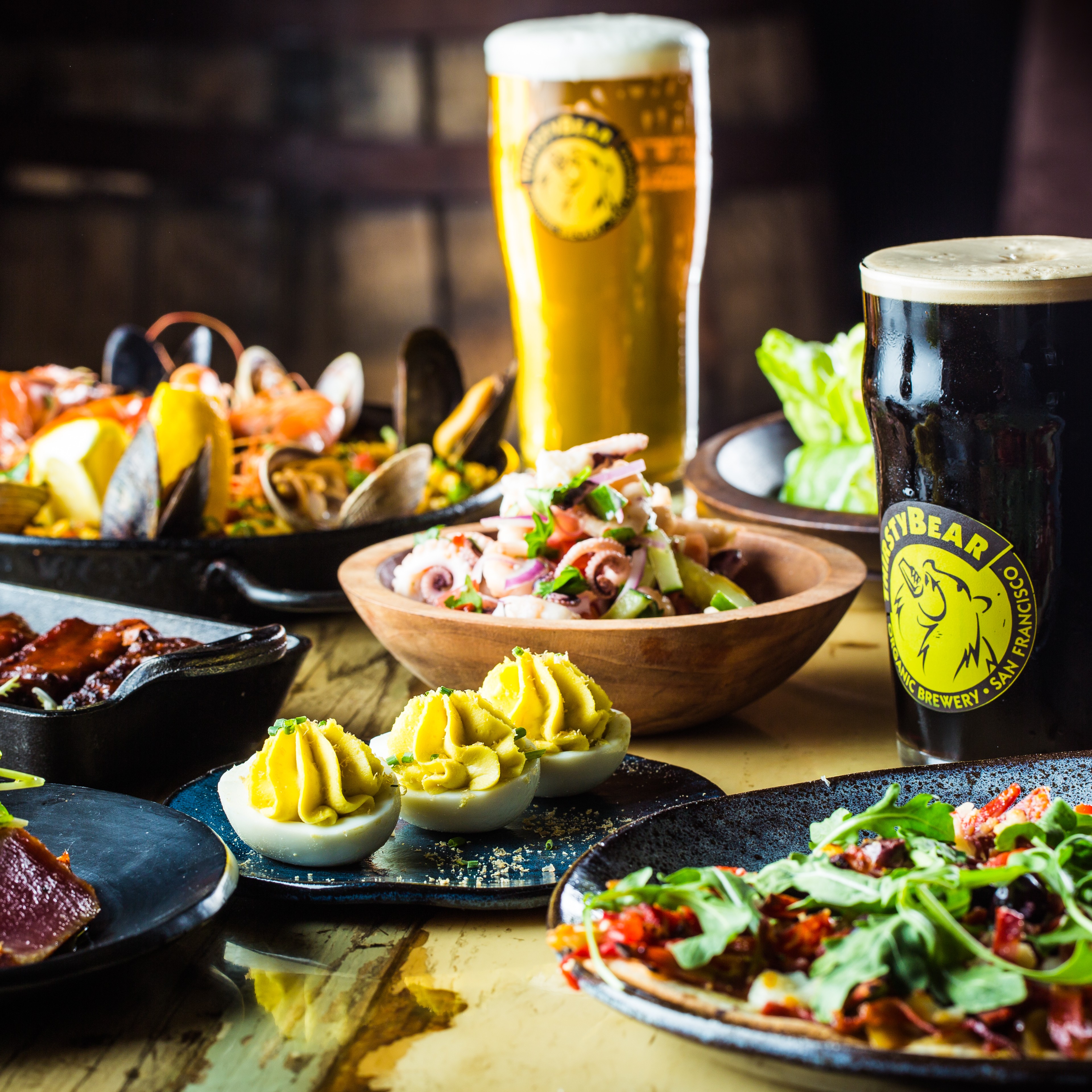 A table with various dishes of food, salas, seared tuna, deviled eggs, and two pints of beer. All drink and food are presented on the table in reusable plates, bowls, and cups