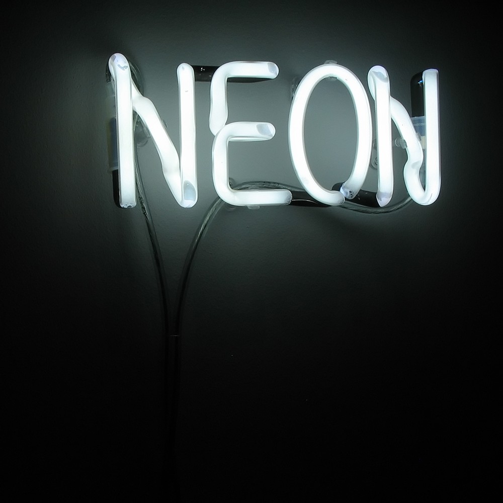 The image shows a neon sign with the word "NEON" lit up in white light. The sign is mounted on a dark wall and is the only source of light in the image, casting a soft glow around the letters. The neon tubes are arranged to spell out "NEON" in all capital letters, with the cabling and mounting structure faintly visible behind the glowing tubes