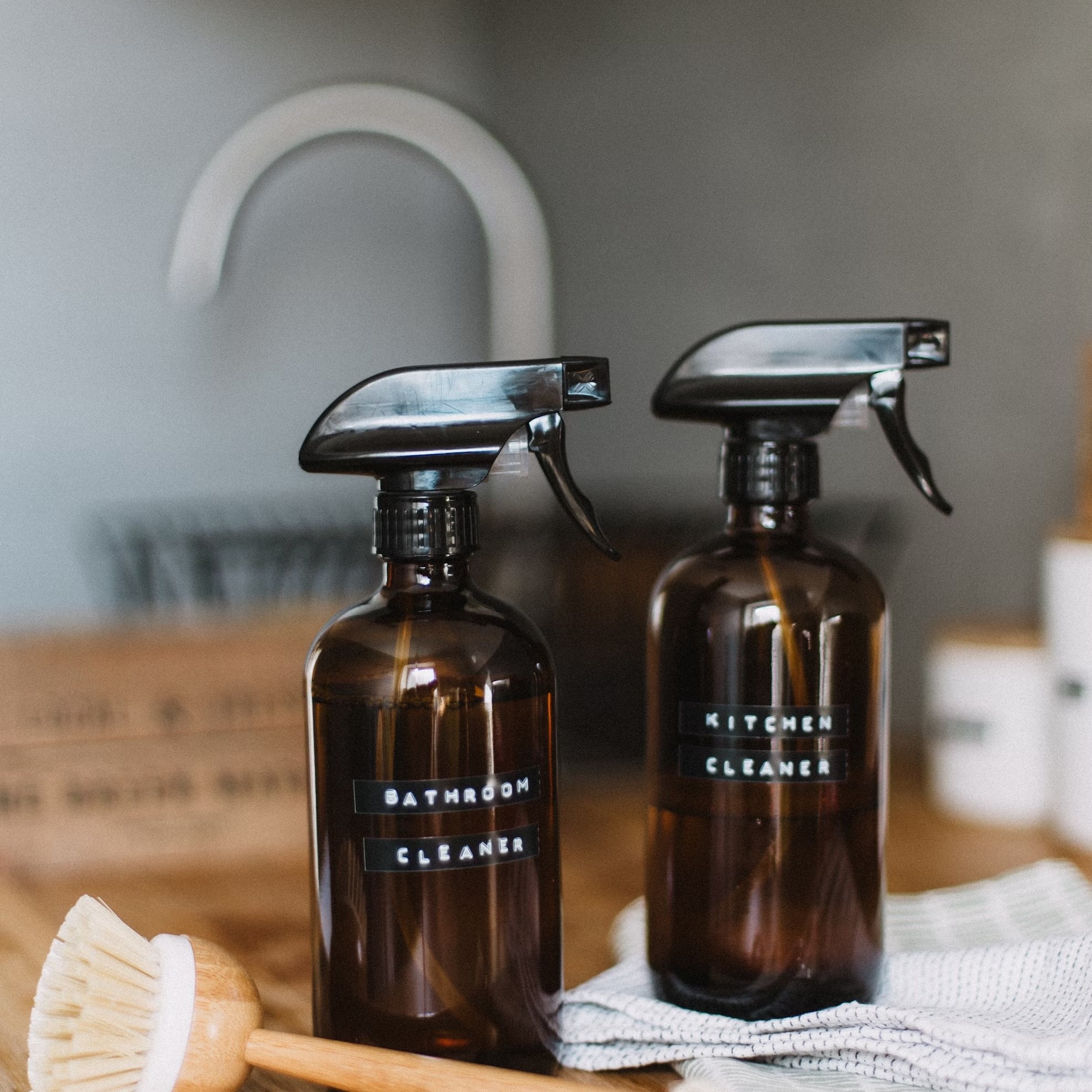 The image shows two brown spray bottles labeled "BATHROOM CLEANER" and "KITCHEN CLEANER," placed on a wooden surface. There is also a wooden dish brush and a white cloth nearby. The setting appears to be a clean and organized kitchen or cleaning area, with a sink and faucet visible in the background. The overall aesthetic is simple and eco-friendly, with a focus on natural materials.