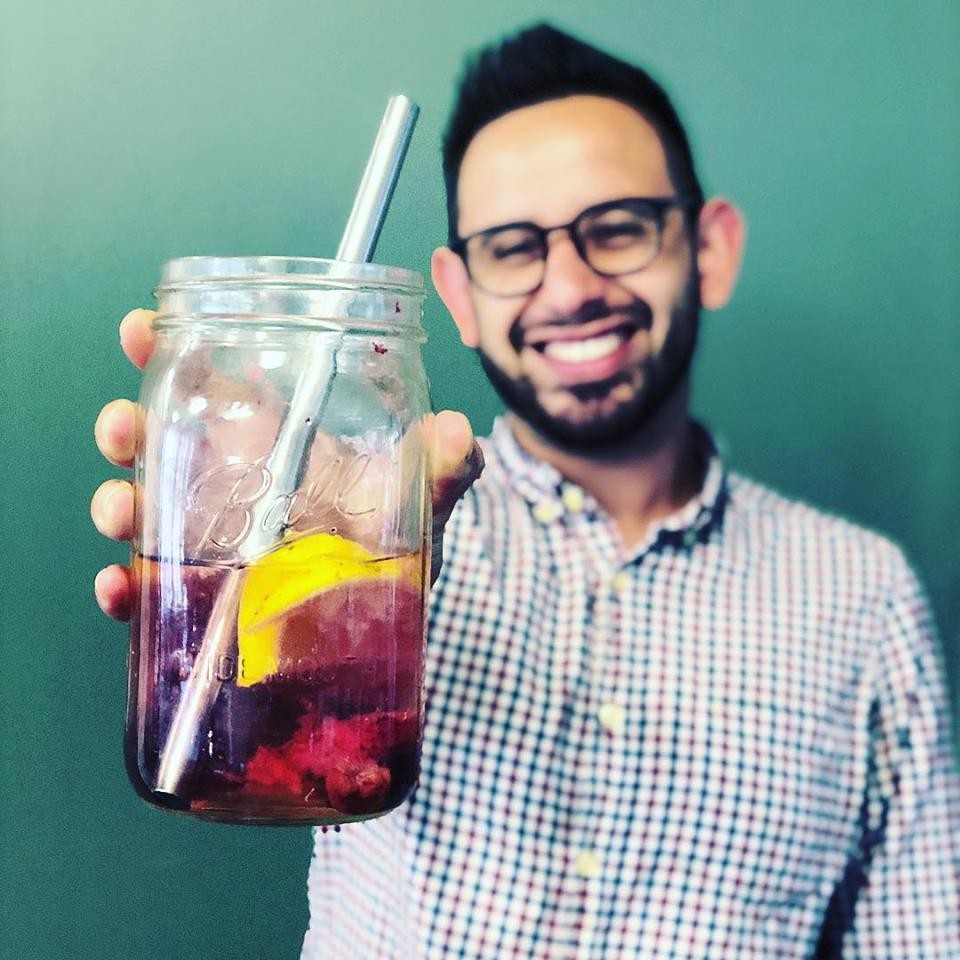 A man with glasses and a checkered shirt is smiling and holding a large mason jar filled with a dark beverage, ice, a metal straw, and a slice of citrus fruit, against a plain green background.