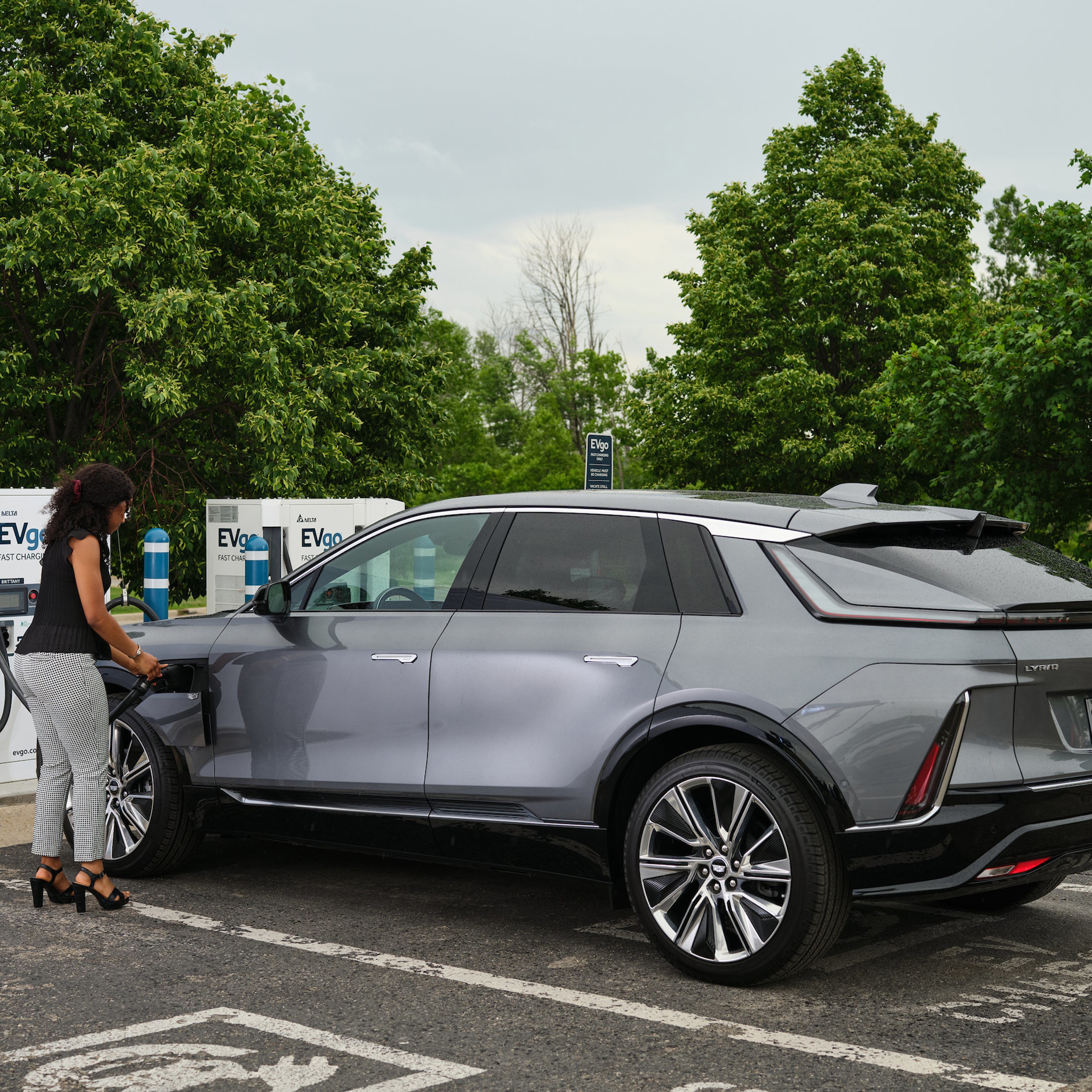 A woman charges a sleek silver electric SUV at an EVgo fast charging station, surrounded by green trees in a parking lot on a cloudy day.