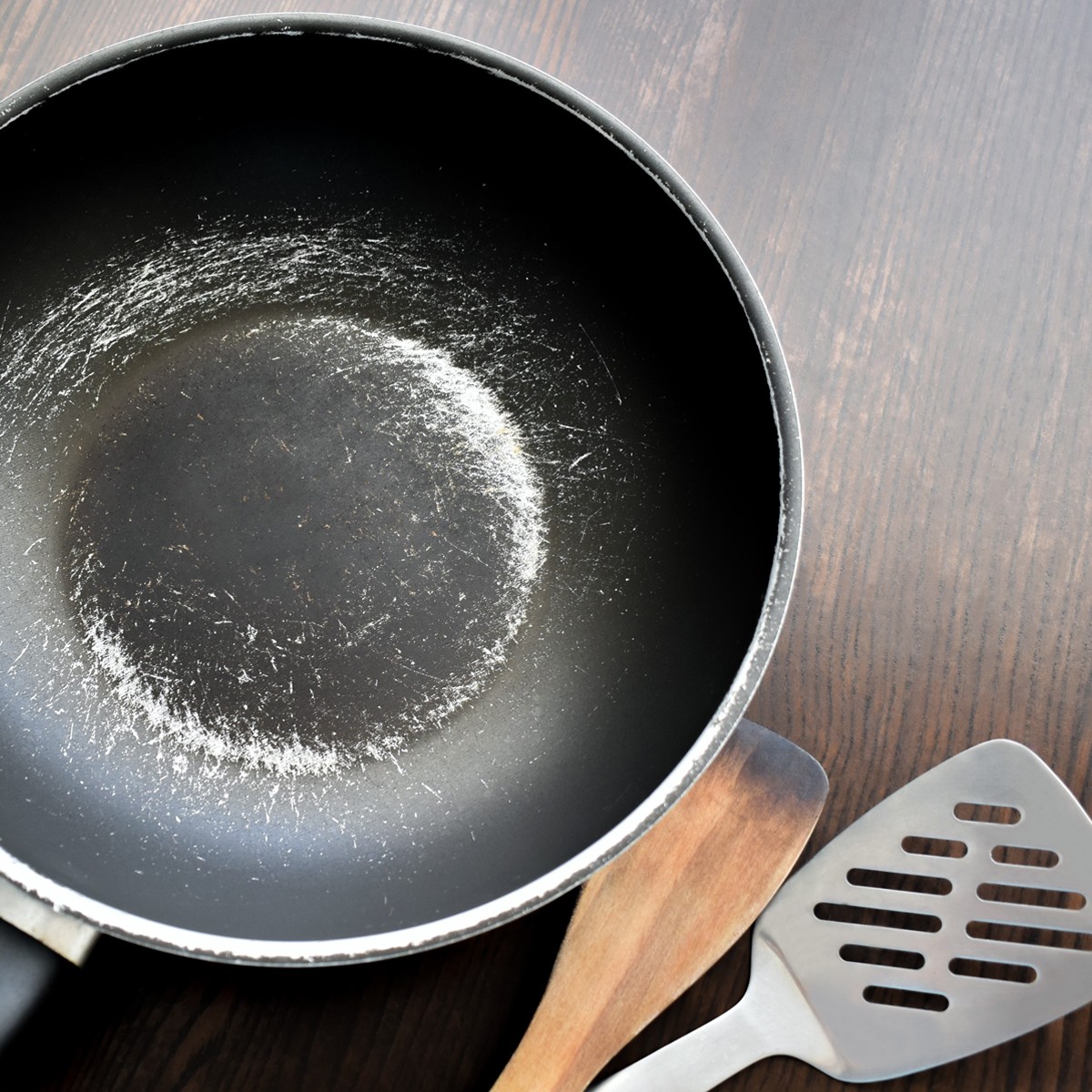 A black frying pan, scratched and worn, sits on a wooden surface. A wooden spatula and a metal slotted spatula are beside it. The interior of the pan shows signs of use, with a circular area of wear in the center.