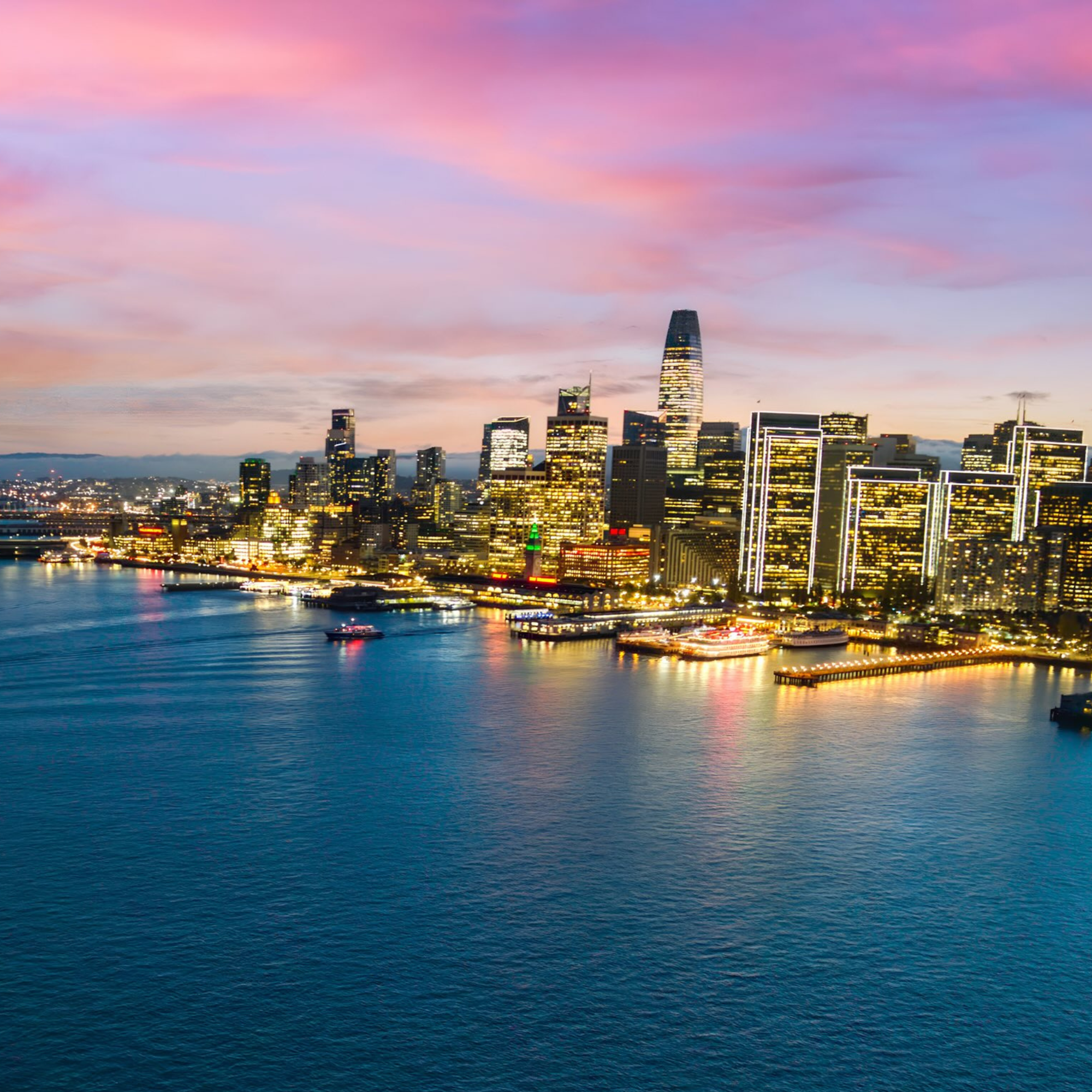 San Francisco skyline at dusk, with the Bay Bridge on the left and illuminated high-rise buildings reflecting on calm bay waters under a pink and purple sunset sky.