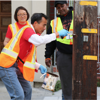 A man wears an orange vest and paints a utility pole brown 