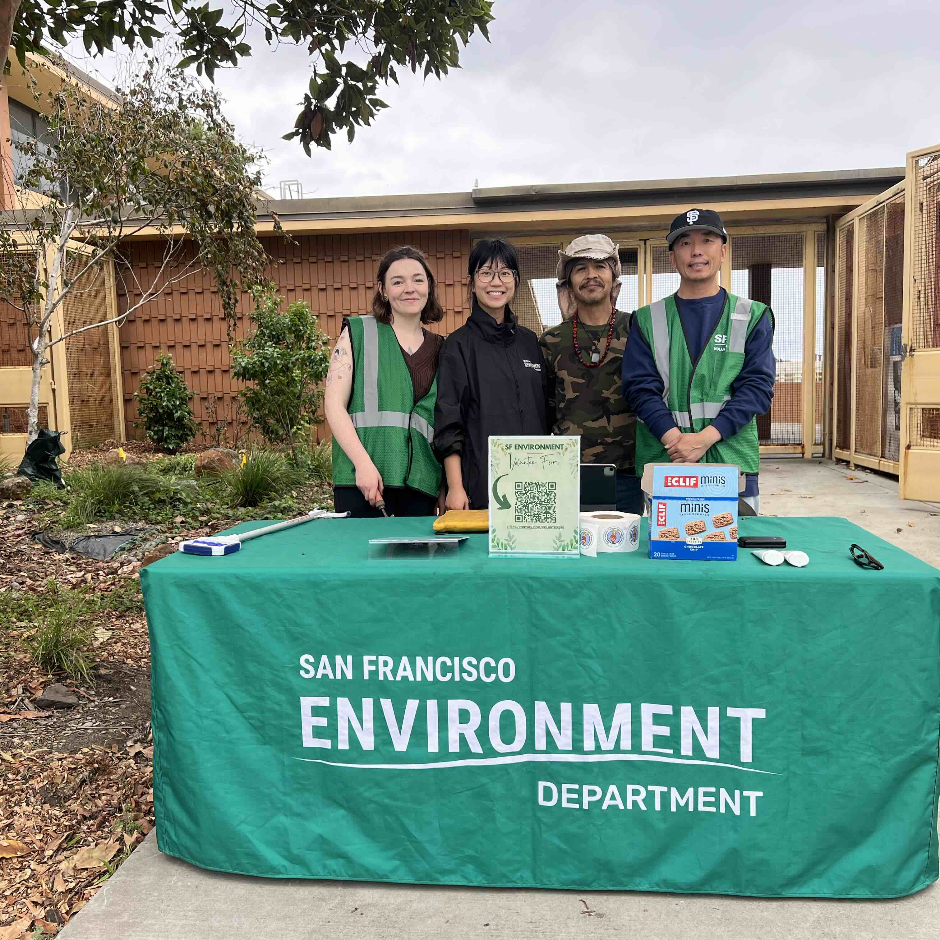 A geren table cover cloth reads SF Environment Department; four volunteers stand behind the table to prepare for an event